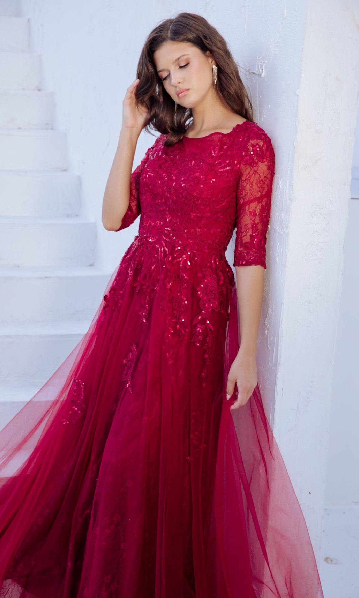 A woman in the Flowy Long Sleeve Long Formal Dress: Eureka 9660, featuring a lace bodice, leans against a white wall by white stairs, looking down with one hand in her hair.